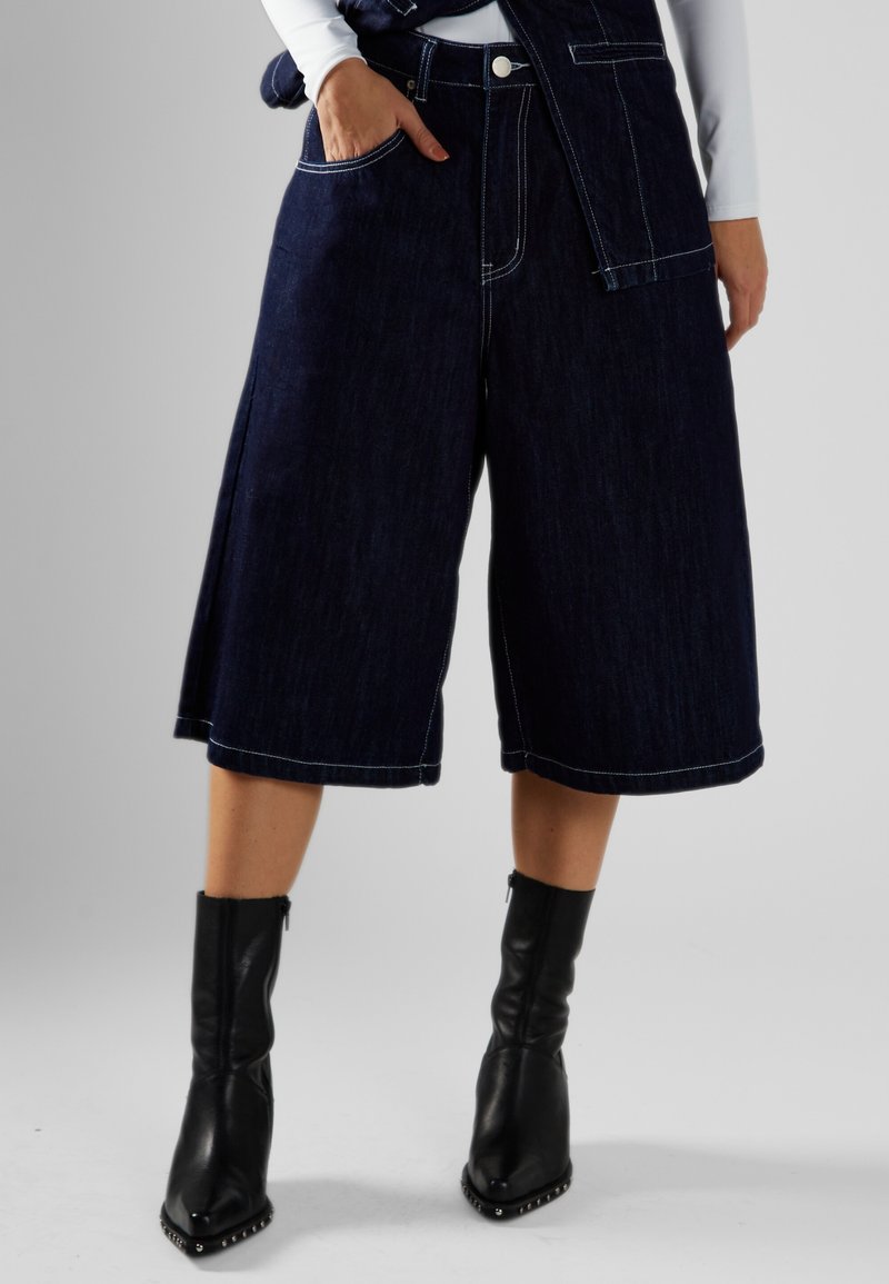 Culottes de mezclilla azul oscuro con costuras blancas contrastantes, bolsillos laterales y un ajuste holgado, combinados con botines negros.