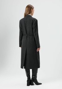 Lange donkergrijze jas met een getailleerde pasvorm, enkele split aan de achterkant en met knopen afgewerkte manchetten, gecombineerd met kniehoge glanzende zwarte laarzen.