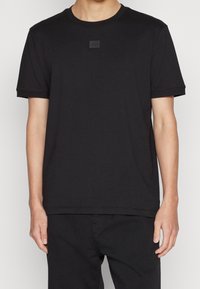 Persona che indossa una maglietta nera a maniche corte senza logo, con un piccolo patch rettangolare nero sul petto, abbinata a pantaloni neri.