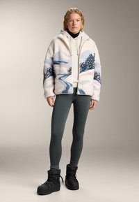 Femeie poartă o jachetă crem din fleece cu model peisaj albastru, hanorac bej, colanți gri închis, șosete gri și bocanci negri de iarnă, stand în fața unui fundal simplu.