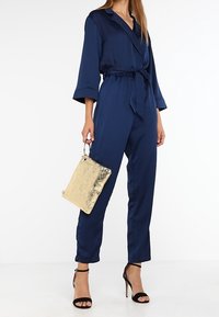 Nawigacyjny satynowy kombinezon z paskiem w talii, szerokimi rękawami i prostymi nogawkami. W zestawie złota metaliczna torebka kopertowa i czarne sandały na cienkich paskach.