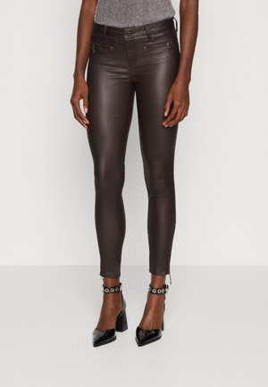 Frau trägt dunkelbraune enge Lederhose mit vorderen Reißverschlusstaschen und schwarze Ankle-Strap-Absatzschuhe aus Lackleder.