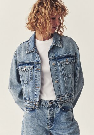 Vrouw met krullend haar, gekleed in een lichtblauwe spijkerjas en bijpassende spijkerbroek, gecombineerd met een eenvoudige witte T-shirt, kijkend naar de zijkant.