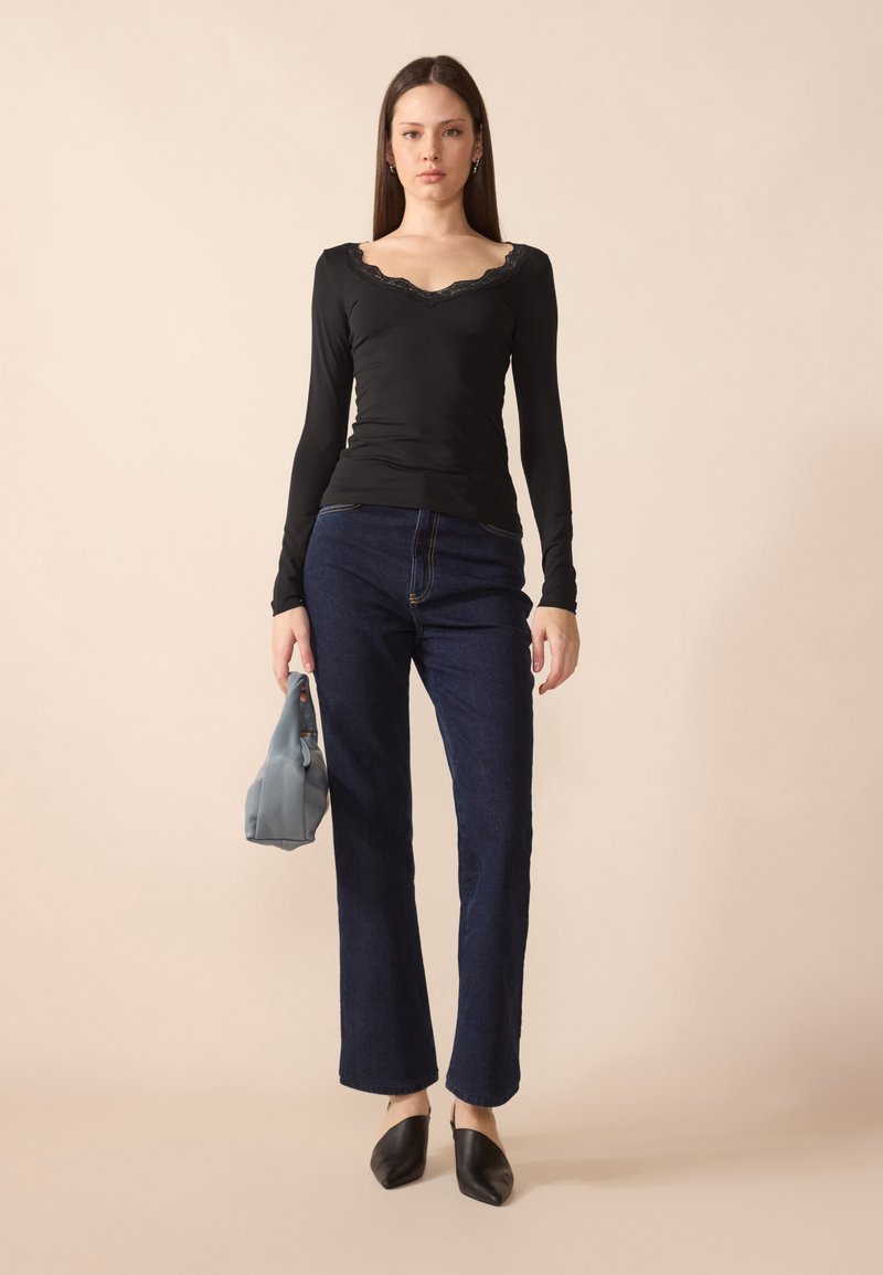 Svart langermet topp med blonder, kombinert med mørkeblå rette jeans og svarte slip-on mules, holder en lyseblå veske.