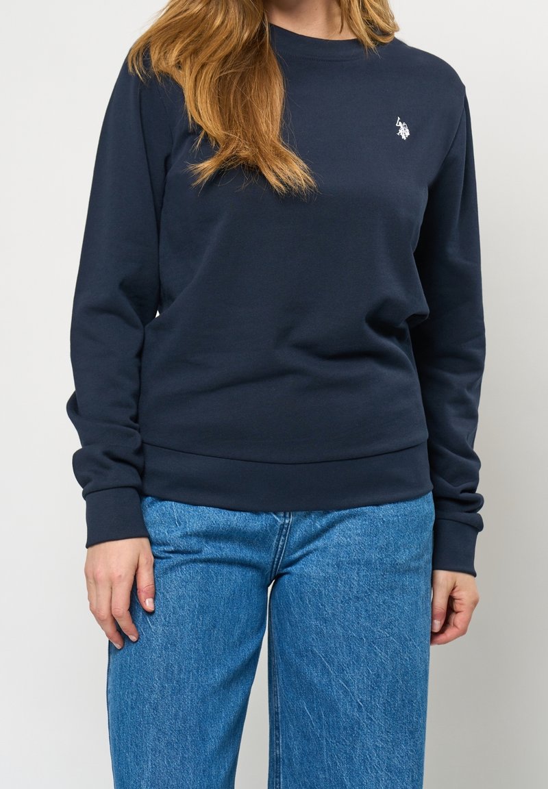 Femme portant un sweat-shirt bleu marine à manches longues avec un petit logo blanc sur la poitrine et un jean bleu, debout devant un fond uni.