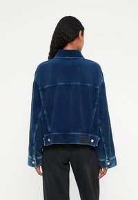 Giacca in denim a coste in blu navy, con un design ampio, collo largo e dettagli con bottoni ai polsini e all'orlo. Superficie strutturata visibile.