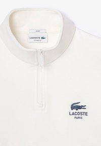 Unisex pulovr se čtvrtinovým zipem v krémové látce, s tmavě modrým logem Lacoste a klasickým střihem. Hladká textura s vysokým límcem a zipovým zapínáním.