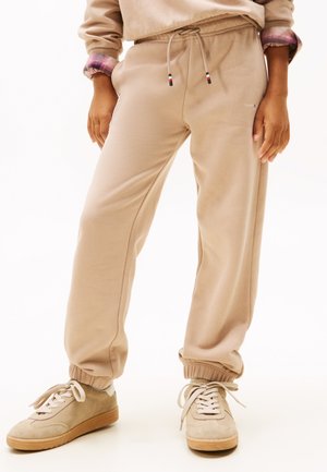 Person, der beige Jogginghosen mit elastischen Bündchen und beigen Sneakern trägt, steht vor einem weißen Hintergrund.