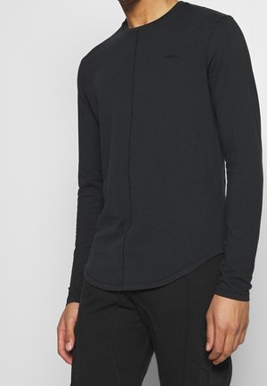 Osoba ubrana w prostą czarną koszulę z długim rękawem z subtelnym przeszyciem z przodu oraz czarne spodnie na neutralnym tle.