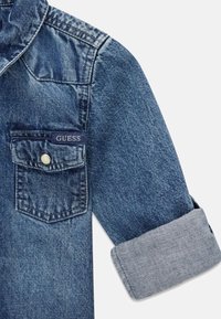 Kurtka dżinsowa w ciemnoniebieskim kolorze, z kieszenią zapinaną na guziki, na której znajduje się niebieska metka z napisem "GUESS", oraz podwiniętymi mankietami z jaśniejszą niebieską podszewką.