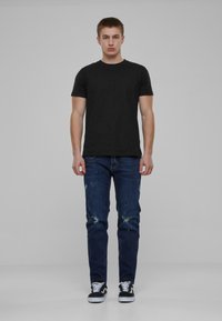 Camiseta de algodón negra, diseño ajustado, combinada con jeans azules desgastados y zapatillas blanco y negro, de pie contra un fondo neutro.