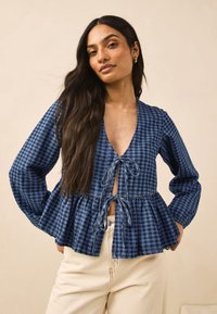 Tamnoplava bluza na karo uzorak s dubokim V-izrezom, zatvaranjem na vezicu, dugim rukavima i proširenim, slojevitim rubom.