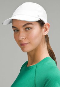 Witte sportieve pet met een gebogen rand en textuurstof, uitgerust met subtiele branding. Model draagt een groene getailleerde shirt.