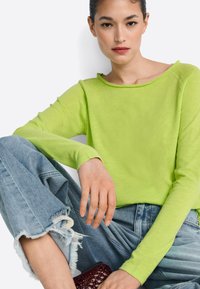 Dlhý rukáv vo farbe limetkovej zelenej s širokým výstrihom a neupravenými okrajmi, v kombinácii s svetlomodrými ošúchanými džínsami s rozstrapkanými manžetami.