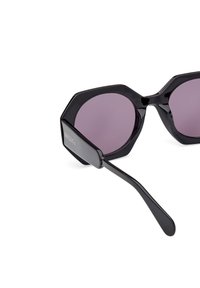 Gafas de sol hexagonales negras con lentes morados oscuros y acabado brillante. Presentan patas negras elegantes con detalles del logo en el costado.