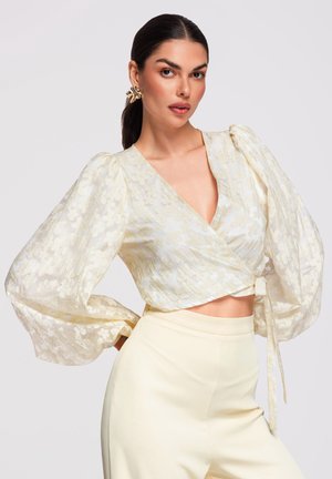 Femme en haut cache-cœur transparent crème à motif floral avec manches bouffantes et pantalon taille haute, posant les mains sur les hanches devant un fond uni.