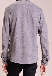 Mand iført langærmet, knaplukket, meleret grå skjorte vendt væk, parret med sorte bukser mod en ensfarvet lys baggrund.