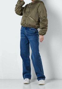 Olivgrön bomberjacka med en mjuk yta, ihopparad med blå raka jeans. Vita sneakers fulländar outfiten.