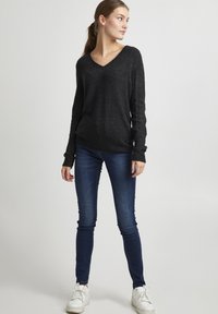 Pull noir à manches longues et col en V en tricot doux et texturé, associé à un jean slim bleu foncé et des baskets blanches.
