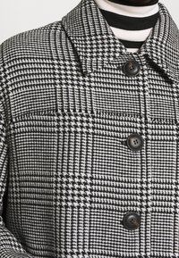 Abrigo de cuadros en blanco y negro con gran cuello, cuatro botones y tela texturizada. Debajo, un suéter de cuello alto con rayas blancas y negras.