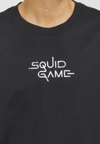 Sort bomulds T-shirt med et hvidt grafisk tryk af teksten "SQUID GAME" i en slidt stil. Rund hals-design, korte ærmer.