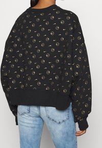 Black sweatshirt with gold circular and Nike logo pattern, ribbed hem, and long sleeves. Worn over light blue jeans.