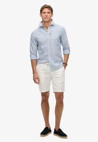 Blau-weiß gestreifte Hemd mit Kragen, weiße Shorts und schwarze Slippers. Modell steht aufrecht, Hände an den Seiten, minimaler Schmuck.