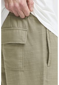 Oliivinvihreät, teksturoidut shortsit, joissa on takataskun yksityiskohta. Rentomalli ja hengittävä kangas. Vähän näkyvissä valkoinen paita vyötäröllä.