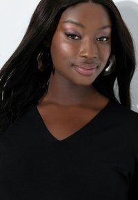 Zwarte V-hals gebreide trui met een gladde textuur, gedragen door een persoon. Grote abstracte zilveren oorbellen completeren de look.