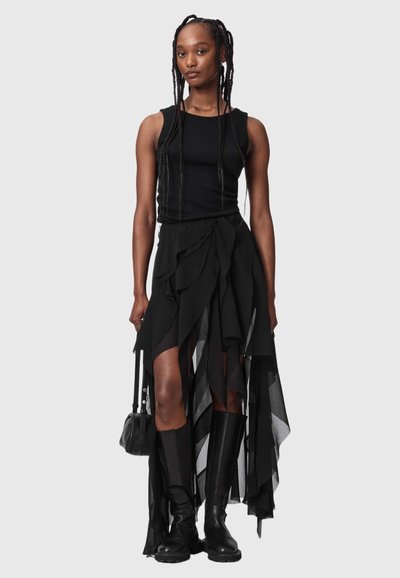 Femme aux longues tresses portant un haut noir sans manches, une jupe noire asymétrique à plusieurs couches, des bottes noires montantes jusqu'aux genoux, et tenant un petit sac à main noir.