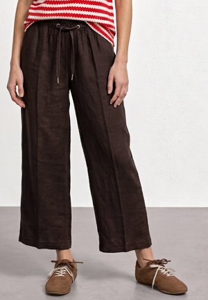 Persoon die losse donkerbruine korte broek draagt met een trekkoord in de taille, een rood-wit gestreept topje en bruine veterschoenen.