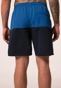 Pantaloni da bagno blu e navy con vita elastica, tasca laterale e blocchi di colore a contrasto. Texture liscia, lunghezza a metà coscia.