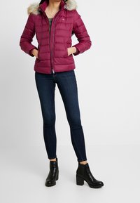 Magenta puchowa kurtka z futrzanym kapturem, z zamkiem błyskawicznym i bocznymi kieszeniami, zestawiona z ciemnoniebieskimi rurkami i czarnymi botkami za kostkę.