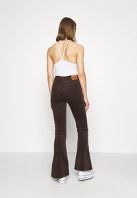 Femme debout, portant un haut blanc à bretelles, un pantalon évasé marron foncé avec l'étiquette Lee, et des baskets compensées blanches, sur fond blanc uni.