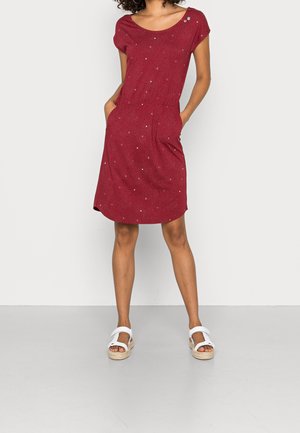 Kvinde iført en knælængde kortærmet rød kjole med små prikmønstre, hænder i lommerne og hvide åbne sandaler med ankelremme.