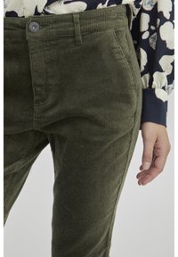Pantaloni in corduroy verde con un motivo testurizzato, dotati di chiusura anteriore con bottone e tasche laterali, abbinati a una camicia a maniche lunghe con motivo.