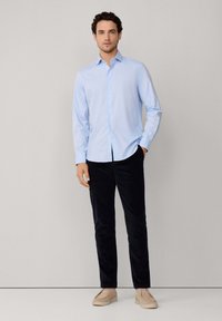 Camisa abotonada azul clara con mangas largas, combinada con pantalones oscuros y zapatos beige. La camisa está hecha de una tela suave, similar al algodón.