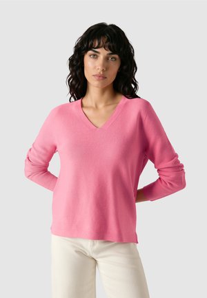 Vrouw met donker krullend haar, gekleed in een roze V-hals trui en lichtgekleurde broek, staand met handen achter haar rug.