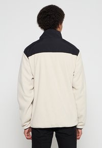 Fleece bunda s krémovým tělem a černými texturovanými rameny. Vysoký límec, dlouhé rukávy a volný střih. Měkká textura po celém povrchu.