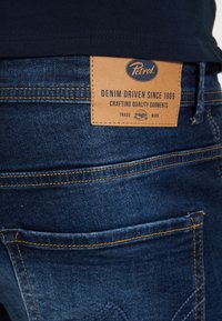 Vaqueros de mezclilla azul oscuro con un parche de cuero que lleva el logotipo "Petrol" y la frase "DENIM DRIVEN SINCE 1989". Incluye costuras visibles y detalles en los bolsillos.