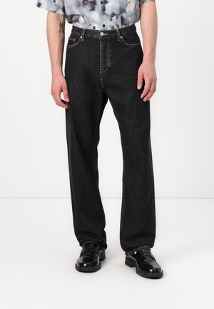 Jean droit noir à taille haute, en denim. Présente des coutures visibles et un design traditionnel à cinq poches, porté avec des chaussures noires.