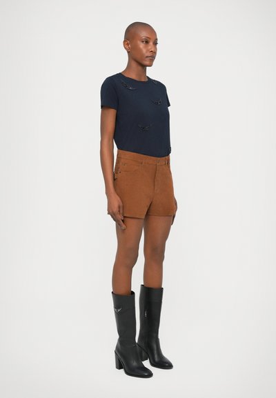 T-shirt marine avec des accents texturés, shorts en daim marron et bottes noires à talons hauts, présentés en pose debout sur fond blanc.