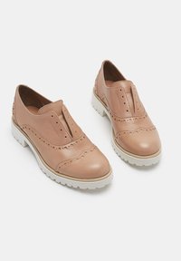 Une paire de chaussures brogues en cuir beige à enfiler avec des semelles en caoutchouc texturé blanc sur un fond blanc.