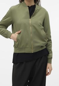 Kvinna klädd i en olivgrön bomberjacka med gulddragkedja, svart lager-på-lager topp och svarta byxor, med handen i fickan på jackan.