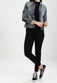 Osoba ubrana w wyblakłą dżinsową kurtkę na czarnej bluzce, czarnych spodniach i czarno-białych sneakersach, stoi z nogami skrzyżowanymi i dłońmi złożonymi.