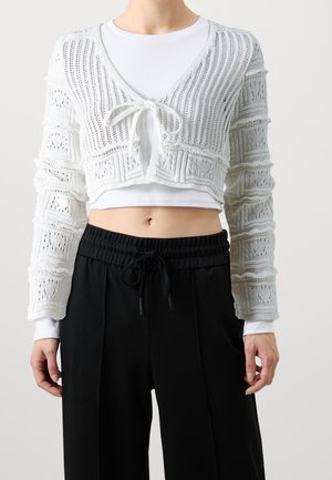 Vrouw draagt een witte, kortgebreide cardigan met knoopsluiting aan de voorkant over een wit shirt met lange mouwen en zwarte broek met trekkoord.