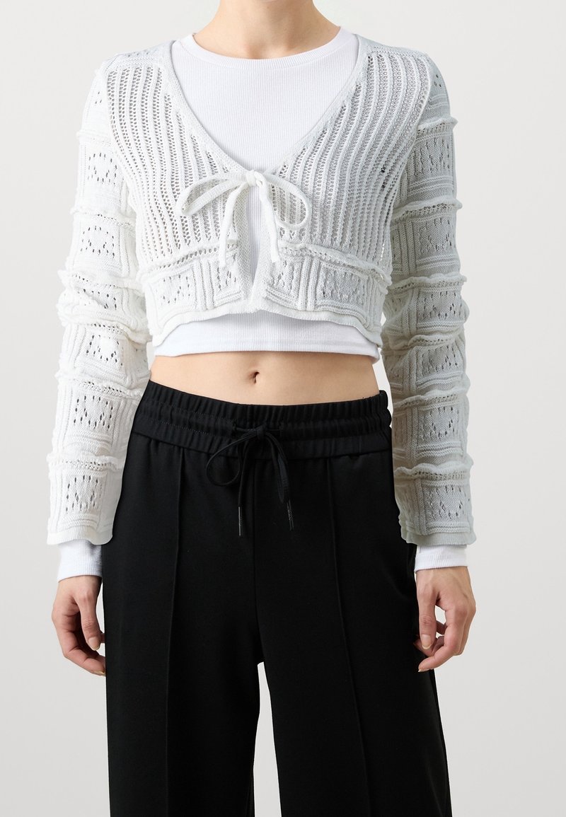 Femme portant un cardigan court en tricot blanc noué à l'avant sur un haut blanc à manches longues avec un pantalon noir à cordon de serrage.