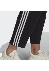 Pantalones de chándal negros con tres rayas blancas a los lados, combinados con zapatillas blancas con detalles en rosa y suelas texturizadas.