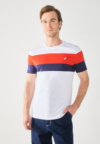 T-shirt en coton blanc avec des rayures horizontales rouges, bleu marine et bleu foncé ; col rond ; petit logo d'oiseau sur la poitrine ; manches courtes.