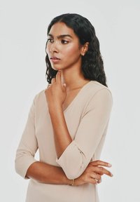 Beżowy, prążkowany top z długim rękawem i dekoltem w kształcie litery V. Modelka ma falujące, ciemne włosy i nosi minimalistyczną biżuterię w kolorze złotym.
